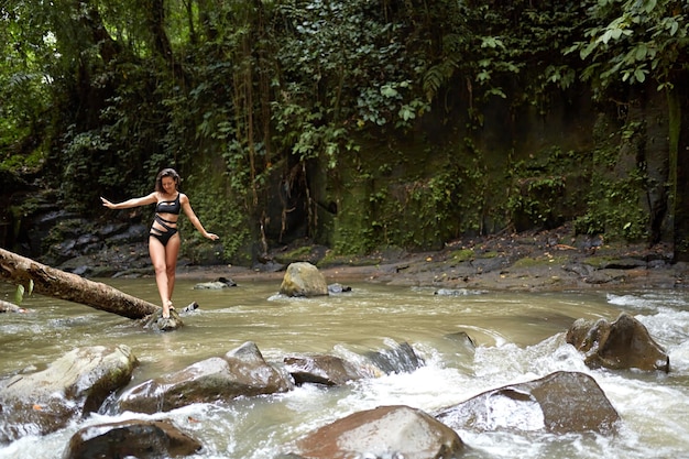 Une jeune fille mince pose sur un tronc d'arbre qui est tombé dans la rivière au milieu d'une jungle dense