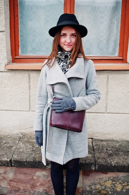 Jeune fille modèle dans un manteau gris et un chapeau noir avec un sac à main en cuir sur les épaules contre la fenêtre de la vieille maison à la rue de la ville.