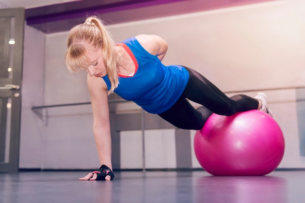 Jeune fille modèle fait des exercices au gymnase. se tenir sur une main. Modèle de fitness blond attrayant effectuant des pompes avec un fitball, mode de vie sain et fitness de concept sportif