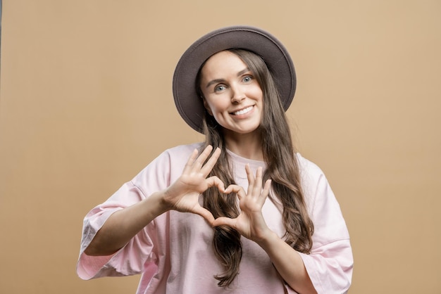 La jeune fille montre un signe de coeur Brunette fille aux cheveux longs portant un chapeau sur un fond beige