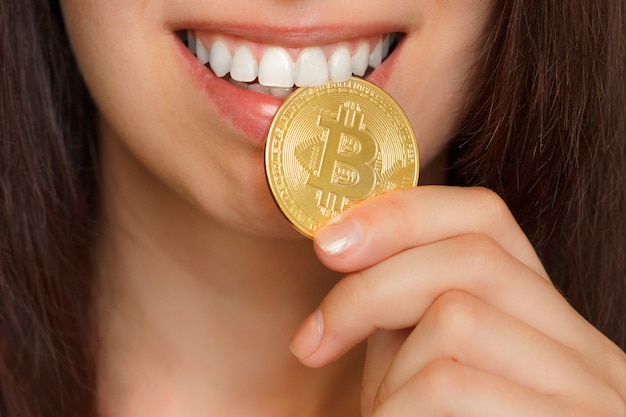 Une jeune fille mord le gros bitcoin BTC doré par des dents blanches. Pièce d'or dans la bouche.