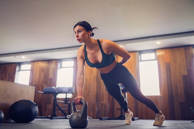 Jeune fille motivée faisant des exercices de planche à l'aide de kettlebells avec une main au gymnase, photo pleine longueur, espace de copie