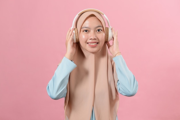 Une jeune fille musulmane souriante écoute sa musique préférée dans des écouteurs blancs, regarde la caméra, s'amuse, mélomane, sur fond rose