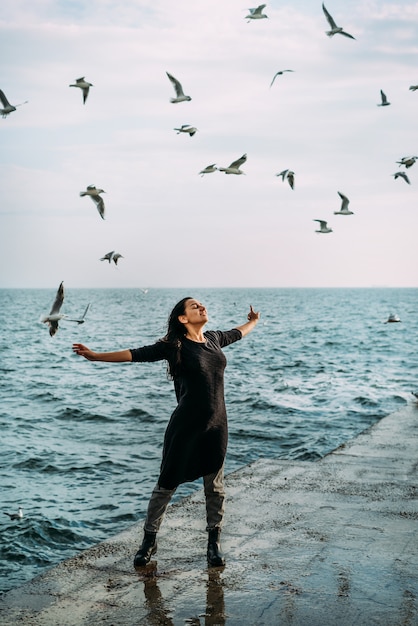 La jeune fille en noir se tient sur la jetée à bras ouverts au soleil et le vent reçoit l'inspiration et la force et la paix intérieure de l'âme