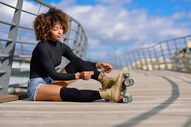 Jeune fille noire souriante assise sur un pont urbain et met sur les patins.