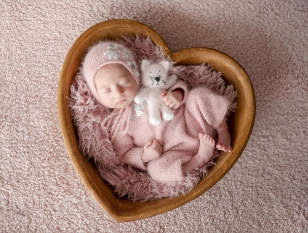 Une jeune fille nouvellement née en costume rose avec un chat jouet dort dans un bol en forme de cœur en bois pendant un nouveau-né professionnel