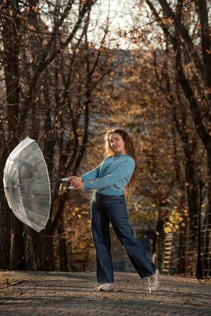 Jeune fille avec un parapluie transparent sourit gentiment Étudiante aux cheveux bouclés pose devant le parc d'automne