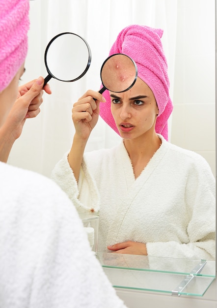 jeune fille à la peau problématique, tenant une loupe sur son acné tout en se regardant dans le miroir