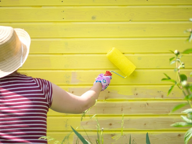 La jeune fille peint un mur en bois avec un rouleau avec de la peinture jaune