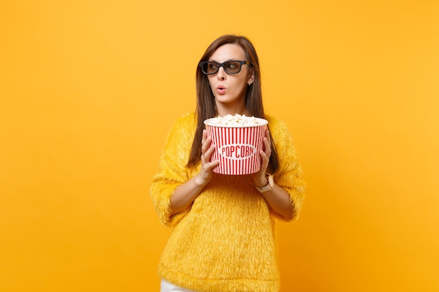 Jeune fille perplexe suspecte dans des lunettes imax 3d regardant un film regardant de côté tenant un seau de pop-corn isolé sur fond jaune vif. Les gens sont des émotions sincères au cinéma, concept de style de vie.