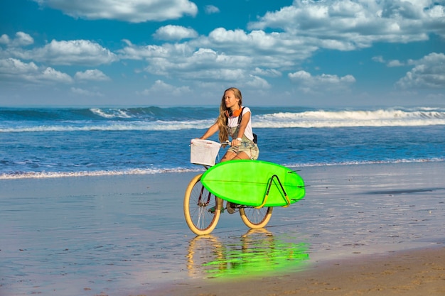 Jeune fille avec planche de surf et vélo sur la plage