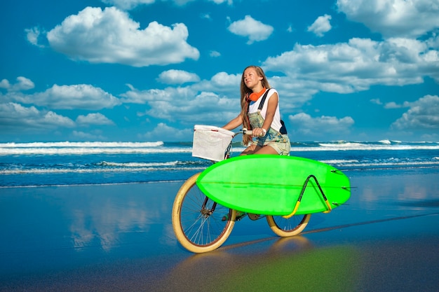 Jeune fille avec planche de surf et vélo sur la plage