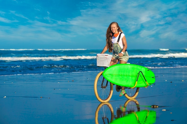 Jeune fille avec planche de surf et vélo sur la plage