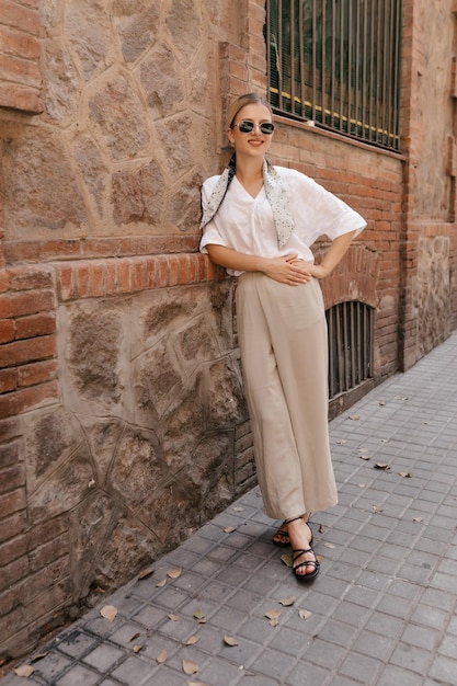 Une jeune fille pleine longueur de bonne humeur se promène dans la ville avec des lunettes de soleil noires. Une jolie blonde