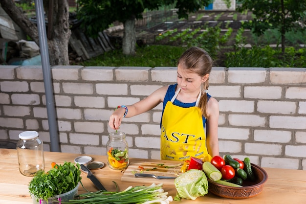 Jeune fille portant un tablier hacher des légumes frais pour la mise en conserve ou la conservation dans des bocaux en verre, à la table d'extérieur près du mur de briques du jardin