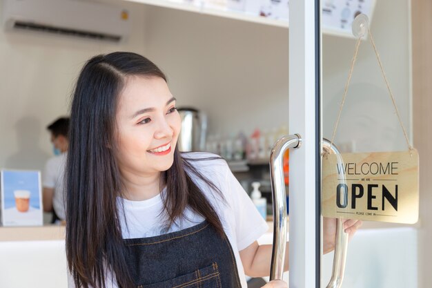 Jeune fille portant un tablier. Happy girl ouvrant sur les portes d'un café et regardant panneau de bois ouvert