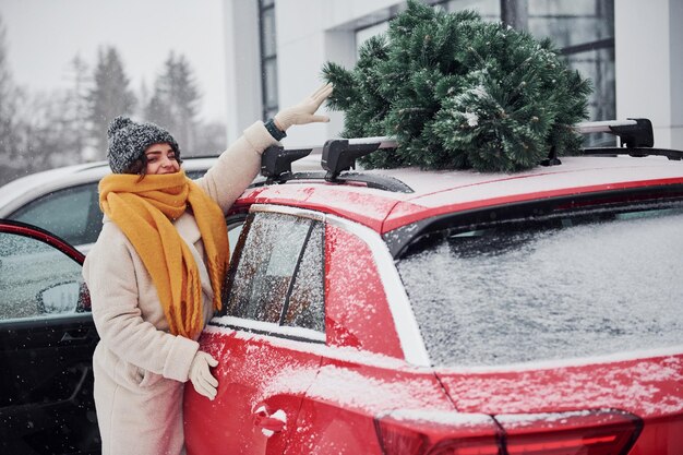 Jeune fille positive debout près de la voiture avec un sapin de Noël vert sur le dessus.