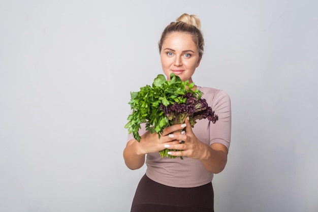 Jeune fille positive donnant un tas d'herbes de cuisine fraîches à la caméra et souriante, légumes verts feuillus, tenant de la laitue d'oseille persillée, une alimentation saine, des aliments biologiques. tourné en studio, fond gris