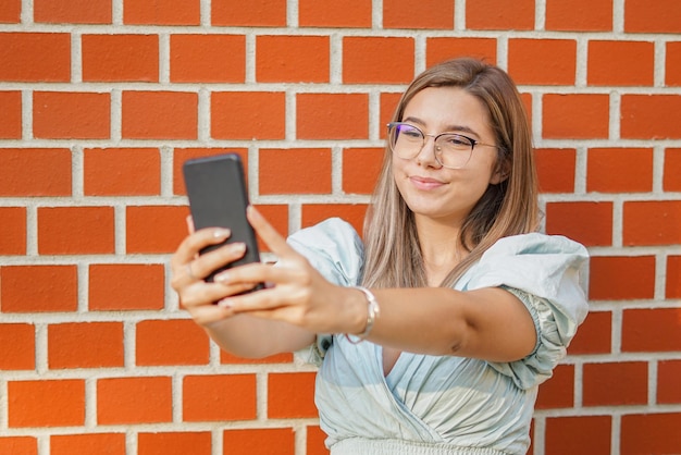 Jeune fille prenant un selfie Souriant à la caméra Bonne étudiante debout près du mur et utilisant son smartphone