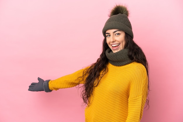 Jeune fille de race blanche avec chapeau d'hiver isolé sur fond violet étendant les mains sur le côté pour inviter à venir