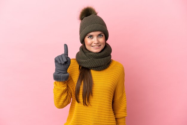 Jeune fille de race blanche avec un chapeau d'hiver isolé sur un mur rose montrant et en soulevant un doigt en signe de la meilleure