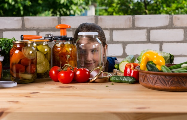 Jeune fille regardant à travers un bocal en verre vide et souriant à la caméra, à l'extérieur dans le jardin à une table en bois recouverte de légumes frais et de pots de conserves