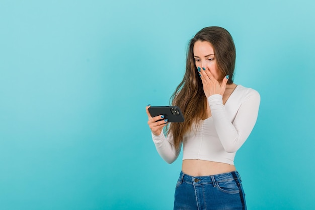 Jeune fille regarde l'écran du smartphone en tenant la main sur la bouche sur fond bleu