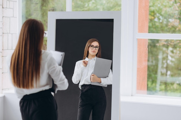 Jeune fille réussie se regarde dans le miroir Fille tenant un ordinateur portable La fille est vêtue d'une chemise blanche et d'un pantalon noir