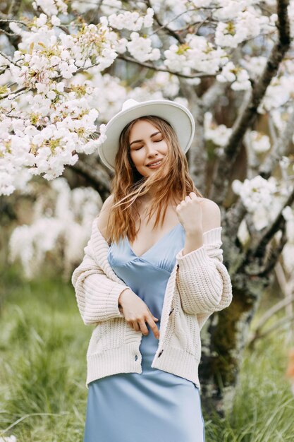 Une jeune fille en robe et un chapeau à la mode se promène en appréciant l'odeur des fleurs en fleurs dans le parc