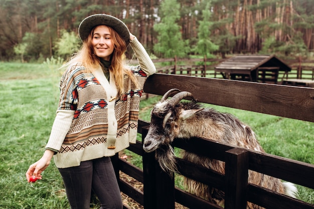 Jeune fille en robe ethnique nourrit le bétail dans une ferme. Une femme en jeans, un pull en laine et un chapeau gris nourrit une chèvre dans un ancien ranch. Contactez le mini zoo en plein air.