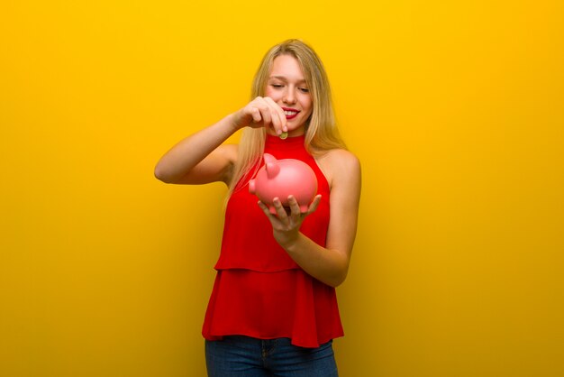 Jeune fille avec une robe rouge sur un mur jaune prenant une tirelire et heureuse parce qu&#39;elle est pleine