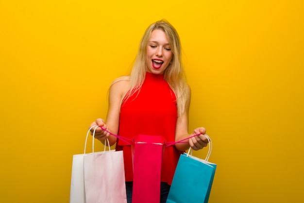 Jeune fille avec une robe rouge sur un mur jaune surpris tout en tenant beaucoup de sacs