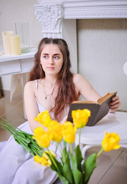 Une jeune fille romantique dans une robe blanche et avec de longs cheveux ondulés lit un livre contre