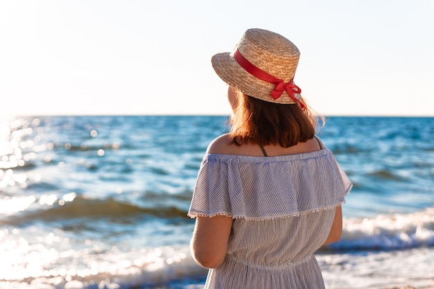 Jeune fille rousse caucasienne solitaire dans un chapeau de paille et une robe d'été avec son dos sur la plage près de la mer