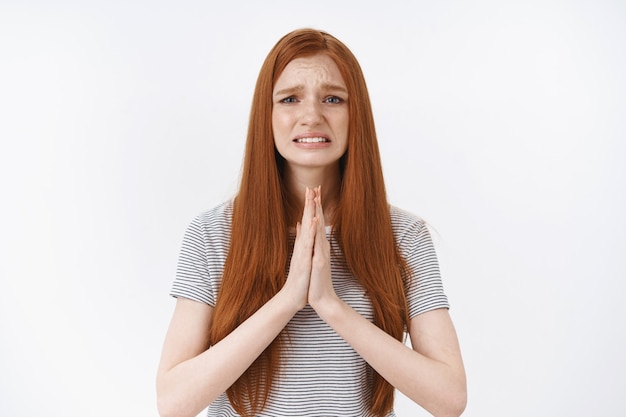 Jeune fille rousse désespérée nerveuse serrer les dents en fronçant les sourcils grimace bouleversante mendier aidez-vous à prendre les mains ensemble prier geste dire s'il vous plaît, plaider faveur, mur blanc debout