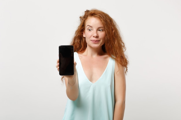 Jeune fille rousse étonnée dans des vêtements légers décontractés posant isolé sur fond blanc, portrait en studio. Concept de mode de vie des gens. Maquette de l'espace de copie. Tenez le téléphone portable avec un écran vide vide.