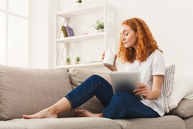 Jeune fille rousse réfléchie lisant sur tablette, assise sur un canapé beige en tenue décontractée. Femme bouclée se reposant à la maison, concept de loisirs et de technologie