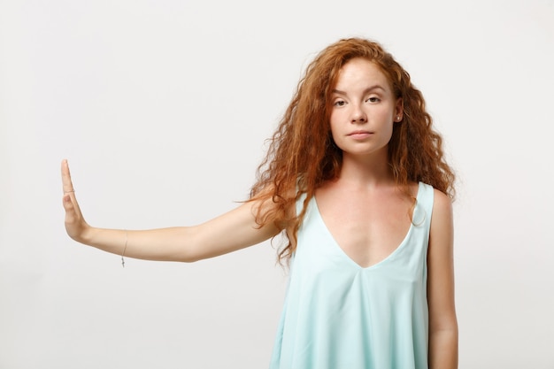 Jeune fille rousse sérieuse dans des vêtements légers décontractés posant isolé sur fond de mur blanc portrait en studio. Concept de mode de vie des gens. Maquette de l'espace de copie. Montrant le geste d'arrêt de côté avec la paume.