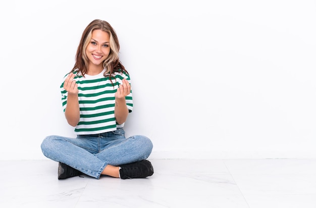 Jeune fille russe assise sur le sol isolé sur fond blanc faisant un geste d'argent