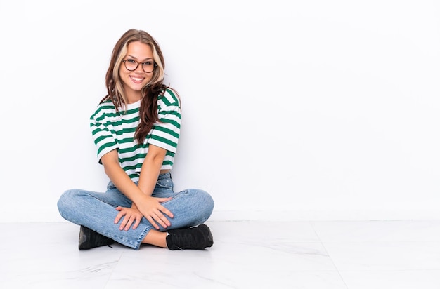 Jeune fille russe assise sur le sol isolé sur fond blanc avec des lunettes et souriant