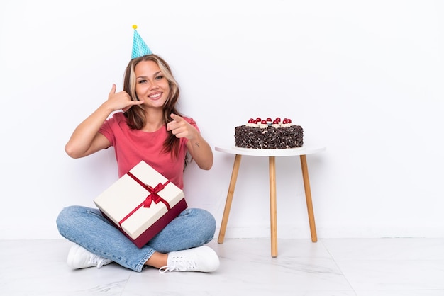 Jeune fille russe célébrant un anniversaire assis sur le sol isolé sur fond blanc faisant un geste de téléphone et pointant vers l'avant