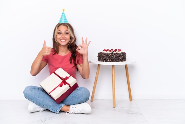 Jeune fille russe célébrant un anniversaire assis sur le sol isolé sur fond blanc montrant le signe ok et le geste du pouce vers le haut