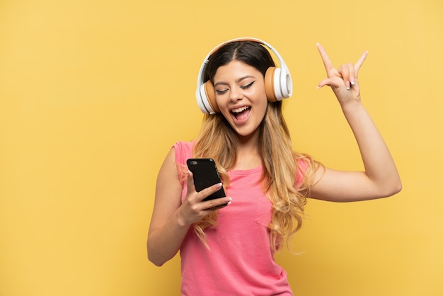 Jeune fille russe isolée sur fond jaune, écoutant de la musique avec un mobile faisant un geste rock