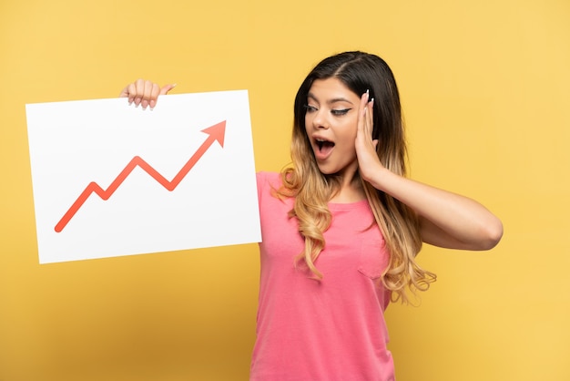 Jeune fille russe isolée sur fond jaune tenant une pancarte avec un symbole de flèche de statistiques croissante avec une expression surprise