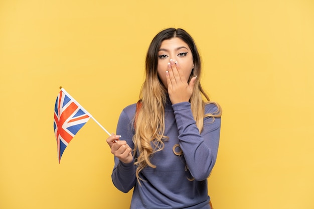 Jeune fille russe tenant un drapeau du Royaume-Uni isolé sur fond jaune heureux et souriant couvrant la bouche avec la main