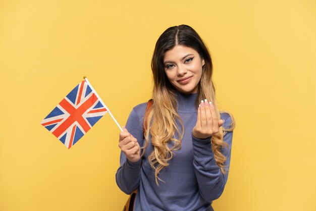 Jeune fille russe tenant un drapeau du Royaume-Uni isolé sur fond jaune invitant à venir avec la main. Heureux que tu sois venu