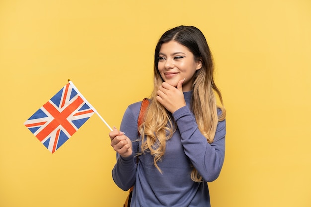Jeune fille russe tenant un drapeau du Royaume-Uni isolé sur fond jaune regardant sur le côté et souriant
