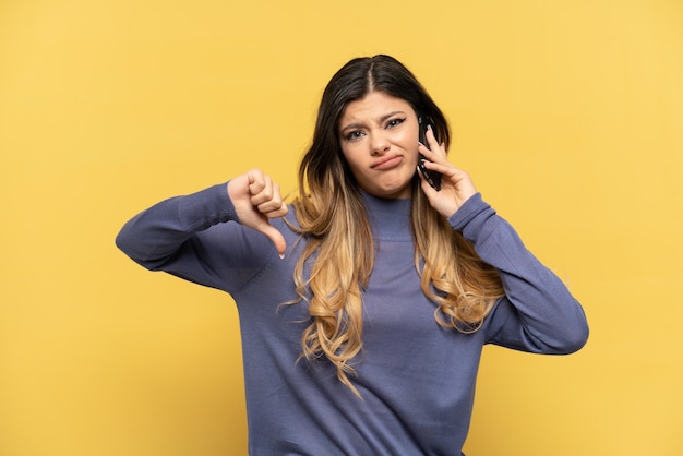 Jeune fille russe utilisant un téléphone portable isolé sur fond jaune montrant le pouce vers le bas avec une expression négative