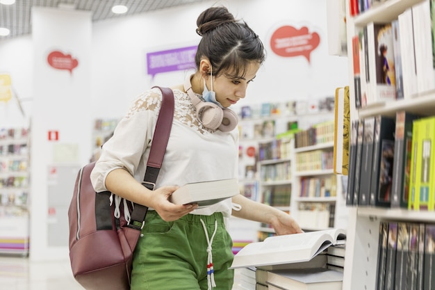 Une jeune fille avec un sac à dos et des écouteurs sélectionne un livre sur les étagères du magasin.