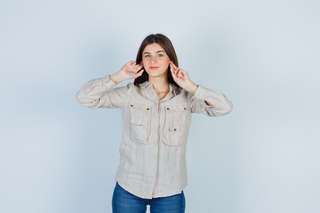 Jeune fille se branchant les oreilles avec l'index en chemise beige, jeans et à la gaieté. vue de face.